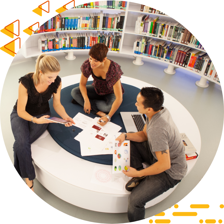 Aerial photo of three students collaborating on 设计 work while seated on a large ottoman, books are in the background.